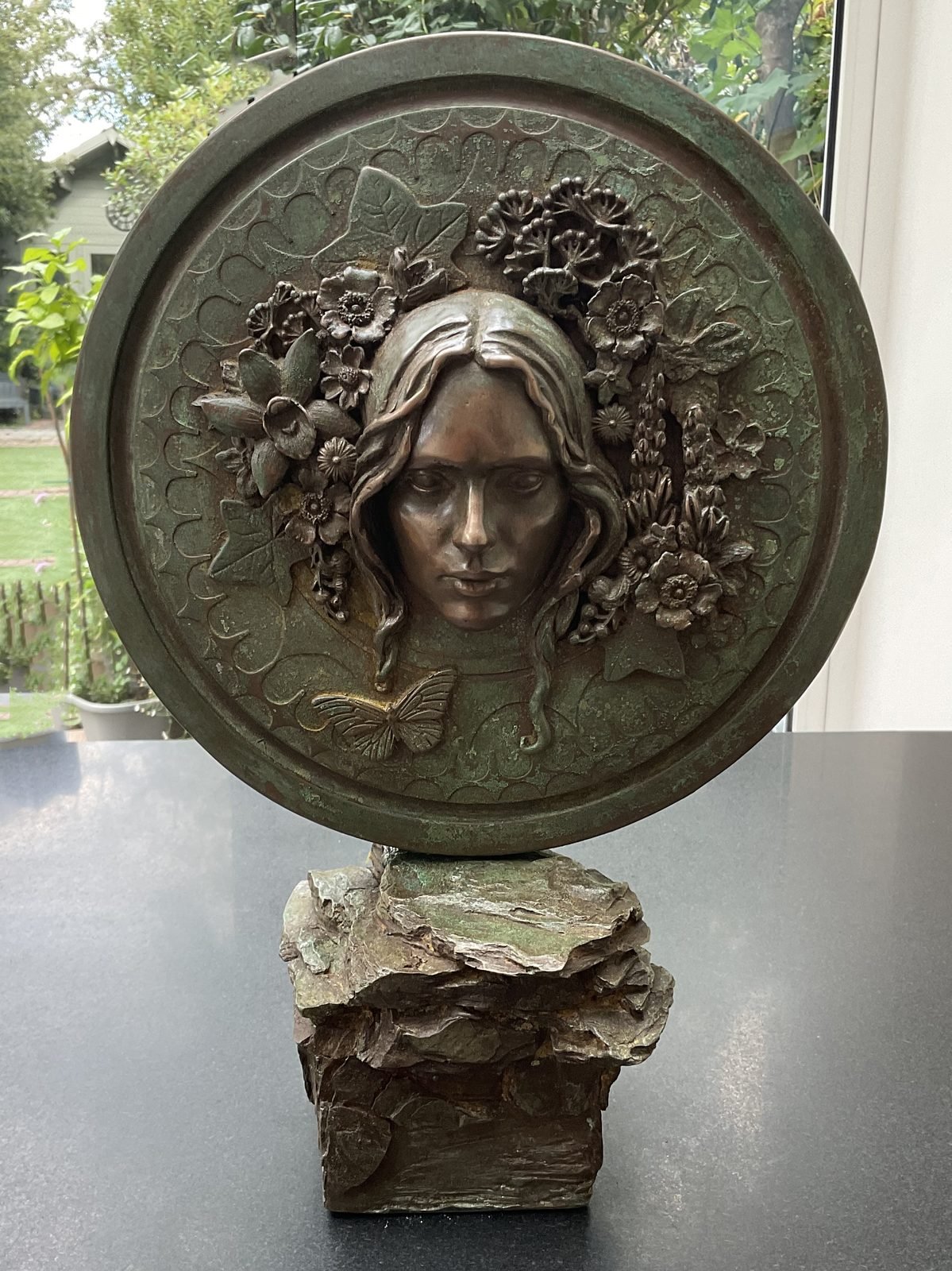 Night And Day: bronze resin roundel, Day side, a bare-headed face framed by poppies, daffodils, lupins and ivy, with a butterfly, on a rough rock base