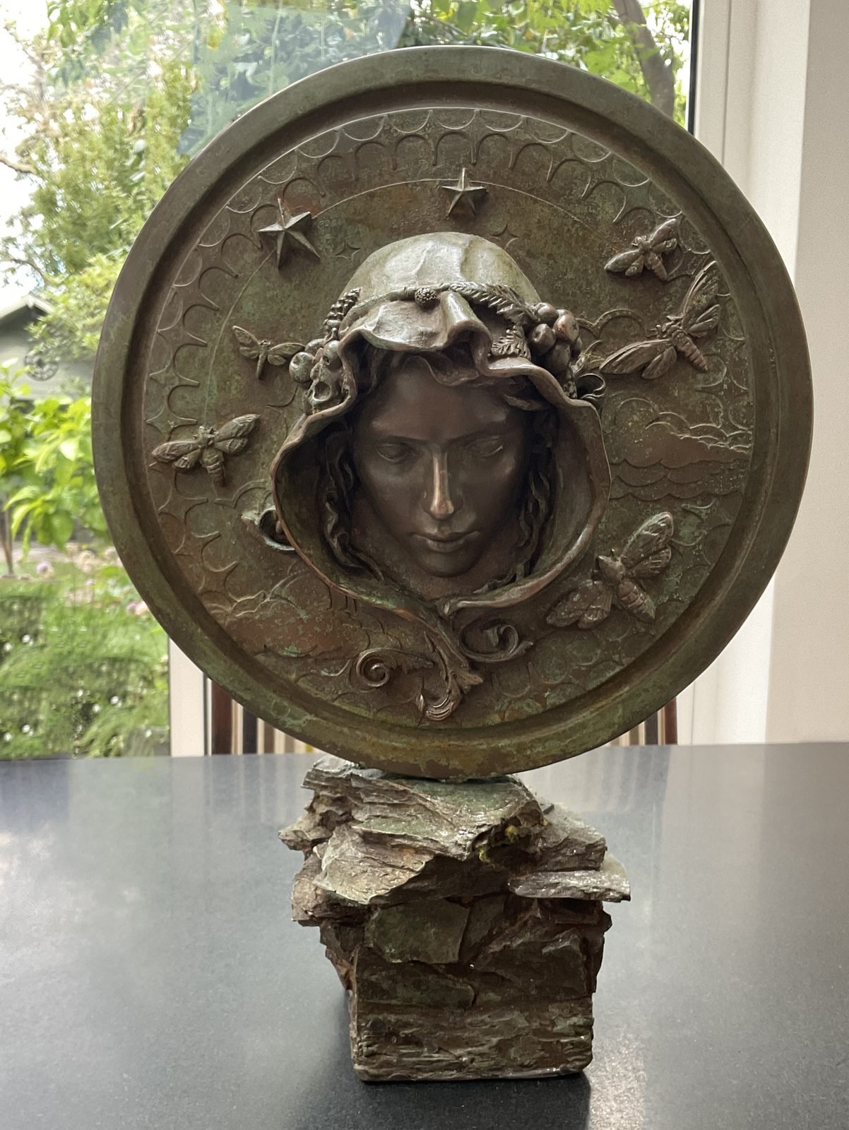 Night And Day: bronze resin roundel, Night side, a hooded face wreathed in grapes and berries, with moths, stars and clouds, on a rough rock base